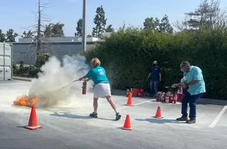 Two people practice using fire extinguishers on a small outdoor fire in a parking lot marked with orange safety cones