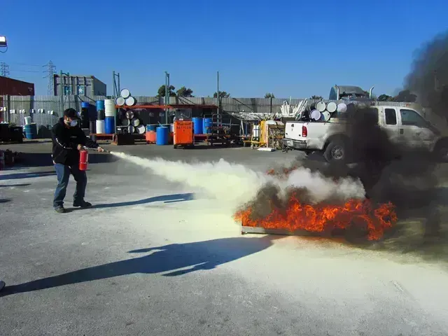 A person uses a fire extinguisher to put out a small fire on asphalt in an industrial area