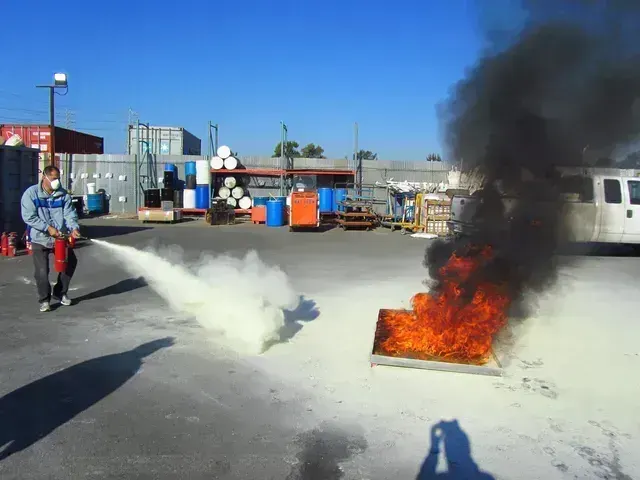A person in a protective mask uses a fire extinguisher to put out a fire in a metal pan outdoors