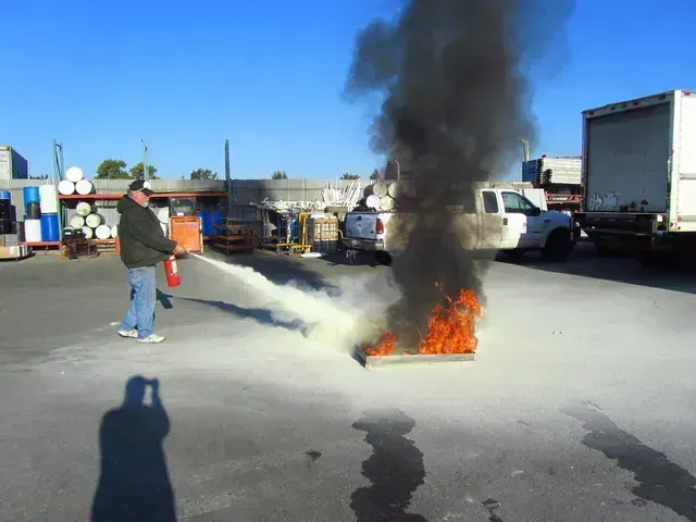 A person wearing a dark jacket and jeans uses a red fire extinguisher to put out a small, intense fire