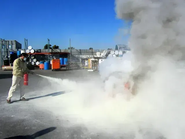 A person in tan work clothes uses a fire extinguisher to spray white powder onto a small fire