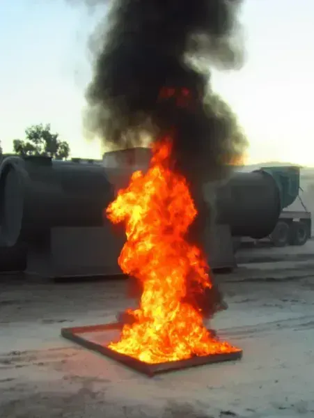 A bright orange fire burns in a rectangular metal tray, producing thick black smoke near industrial tanks