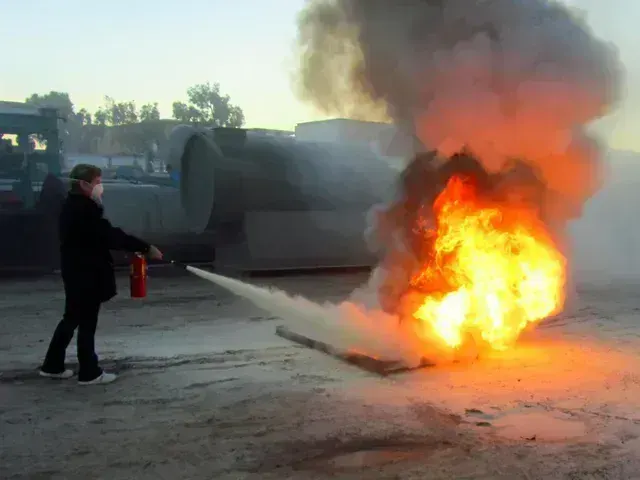 A person uses a fire extinguisher to combat a large, bright orange fire burning on the ground outdoors