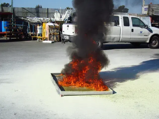 A metal pan of burning fuel emitting flames and thick black smoke in an industrial lot